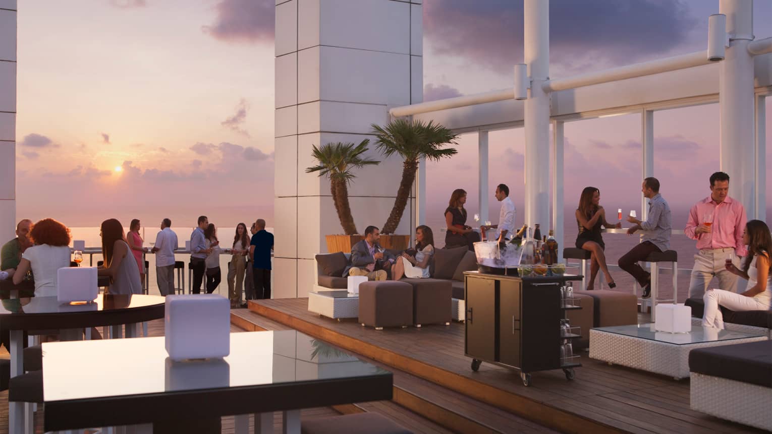 Crowds mingle on rooftop patio on wicker sofas and stand alongside rail as sun sets in background