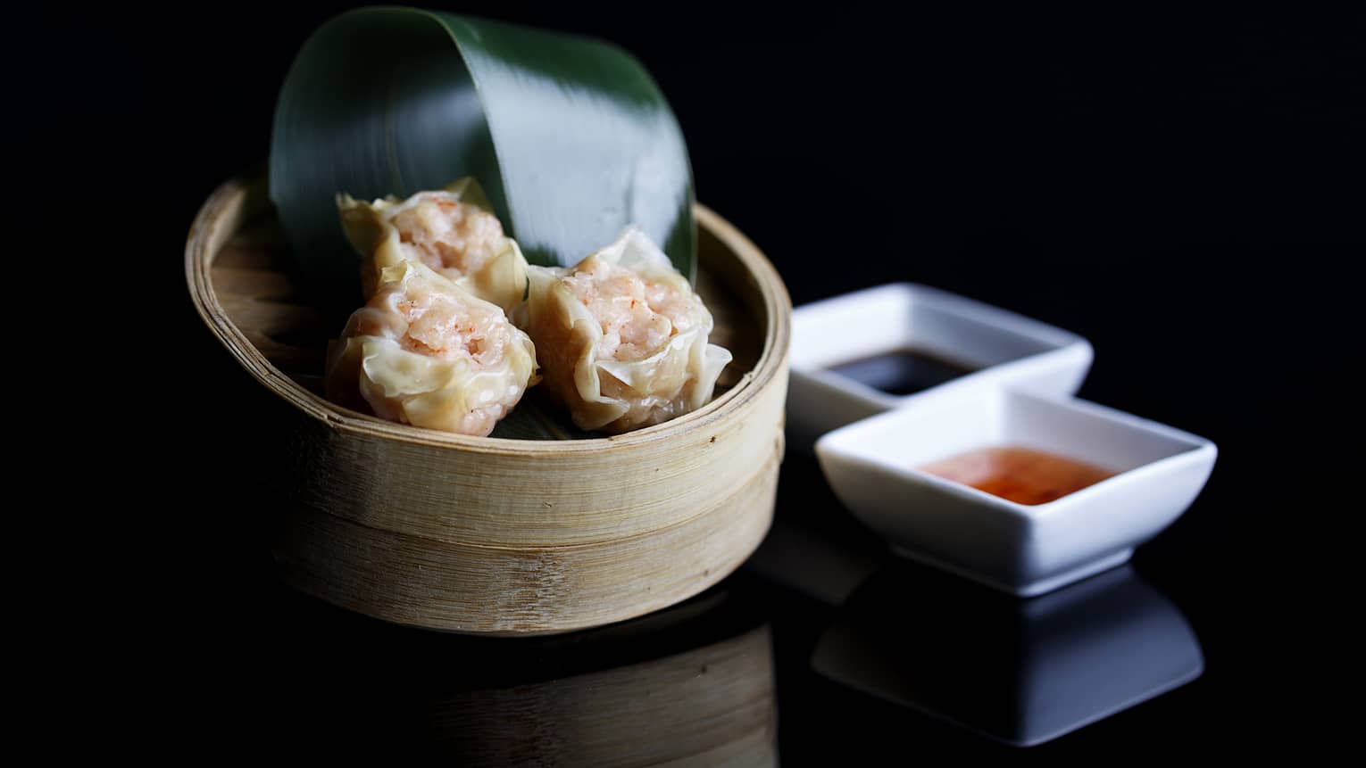 Round bamboo steamer with three homemade shrimp dumplings, banana leaf, two square sauce dishes