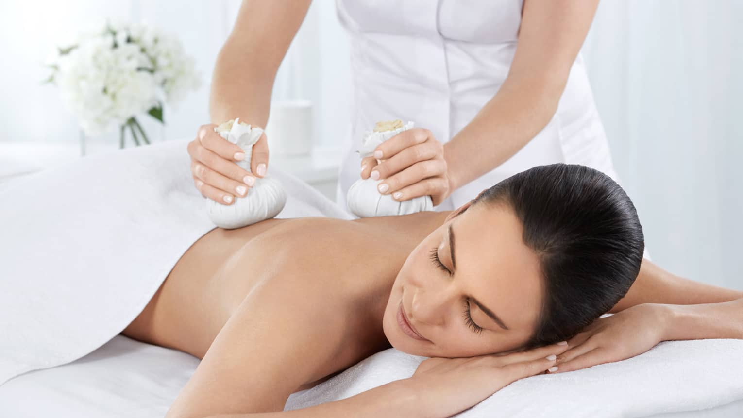 Woman with eyes closed lies under white sheet on massage table, staff presses herbs on shoulders