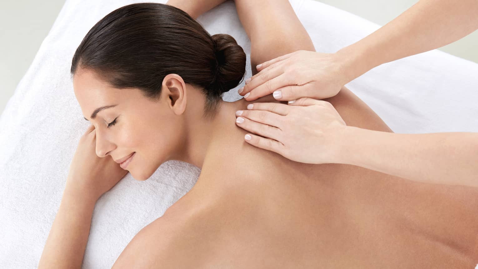 Woman smiles, lies on massage table as hands massage bare shoulders