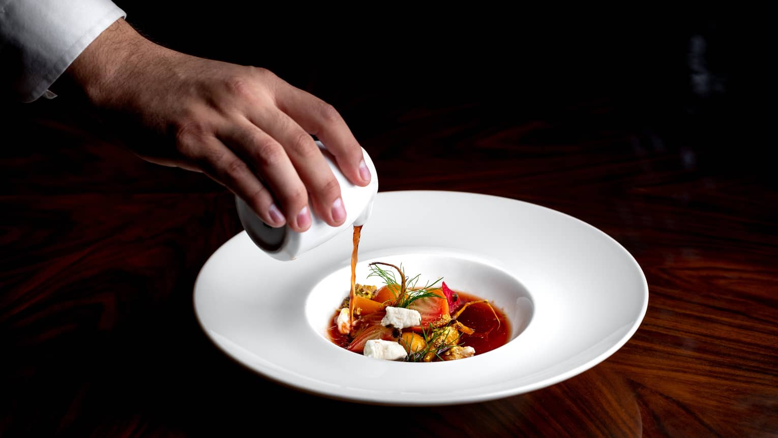 A close-up of a chef pouring a savory broth into a rimmed bowl