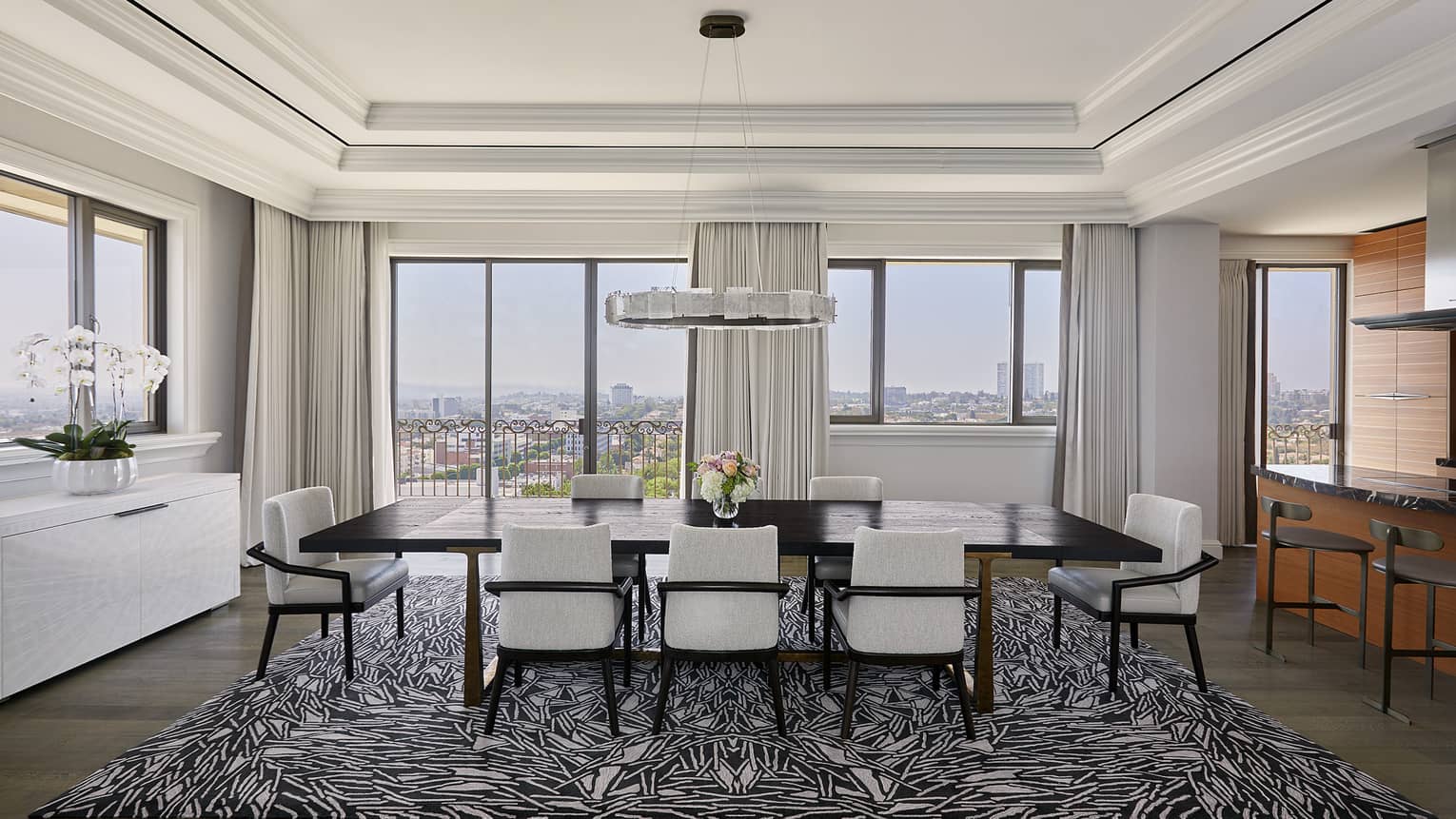 Large dark wood dining table with 10 white chairs, abstract rug, crown molding, hanging light and wall of windows with city view