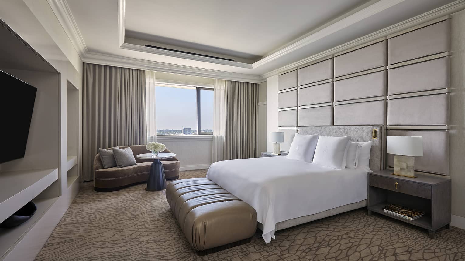 Bedroom with king bed, headboard wall made of up cushioned rectangles, window-side chaise and TV