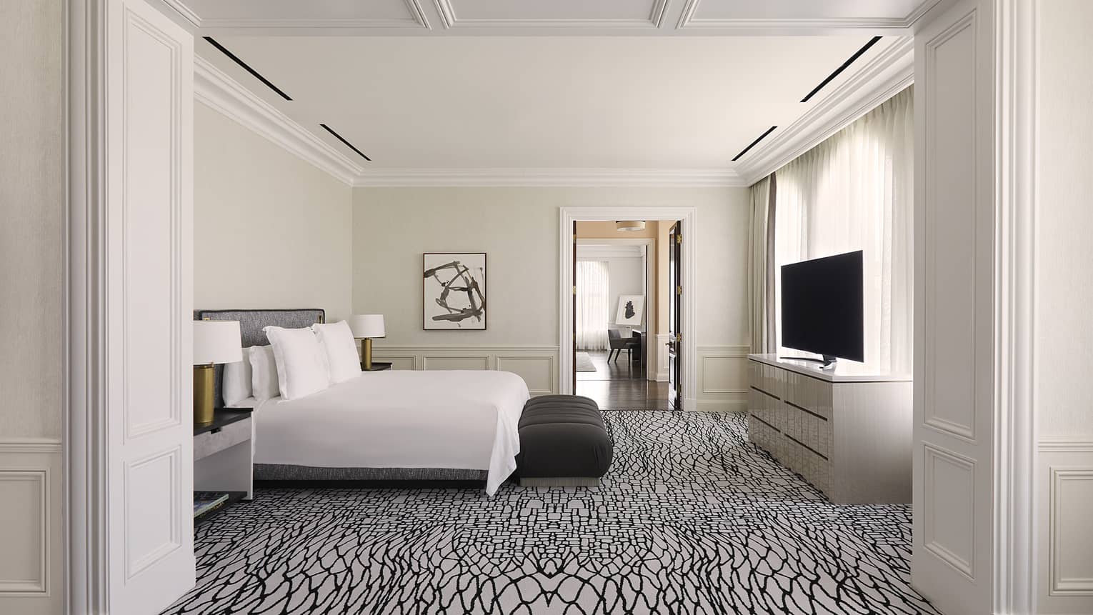 Bedroom with white and black carpeting, white king bed, black bench, TV and dresser