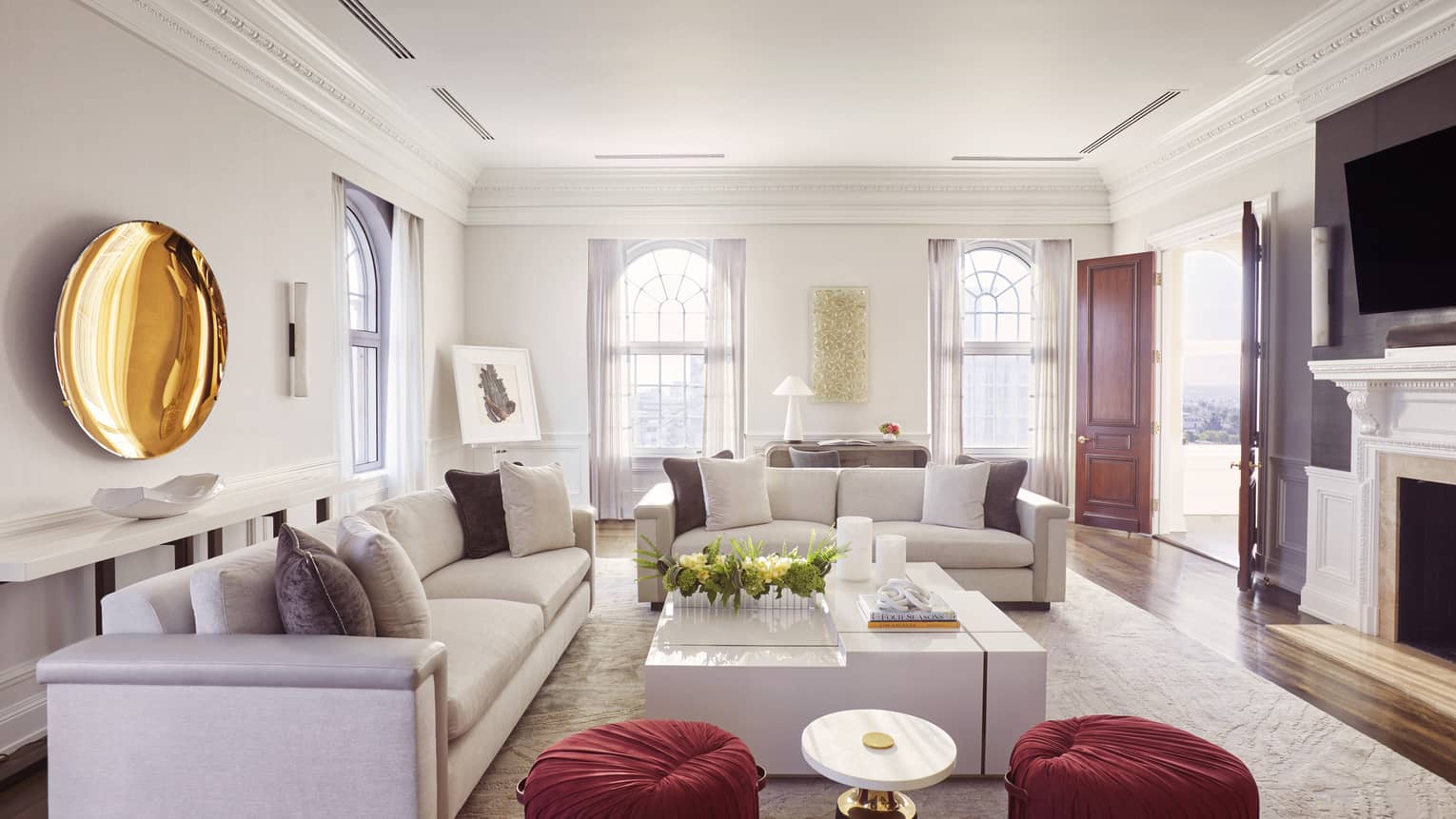 Living room with two sofas, large white square coffee table, two red cushioned stools, gold plate artwork on wall, TV and fireplace