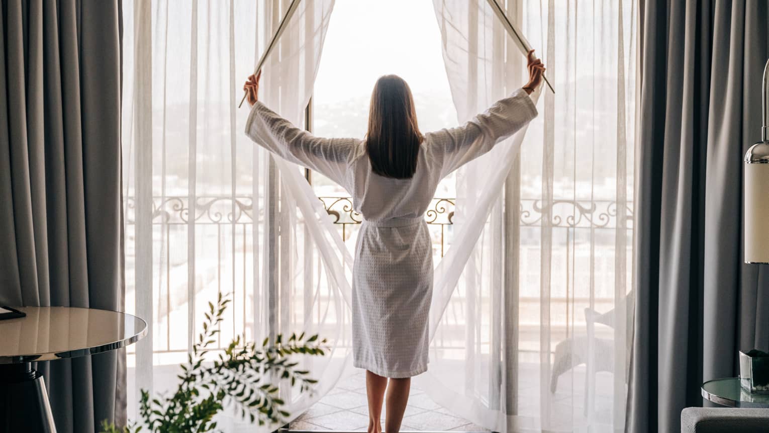 A person standing near a floor-to-ceiling window and opening the curtains