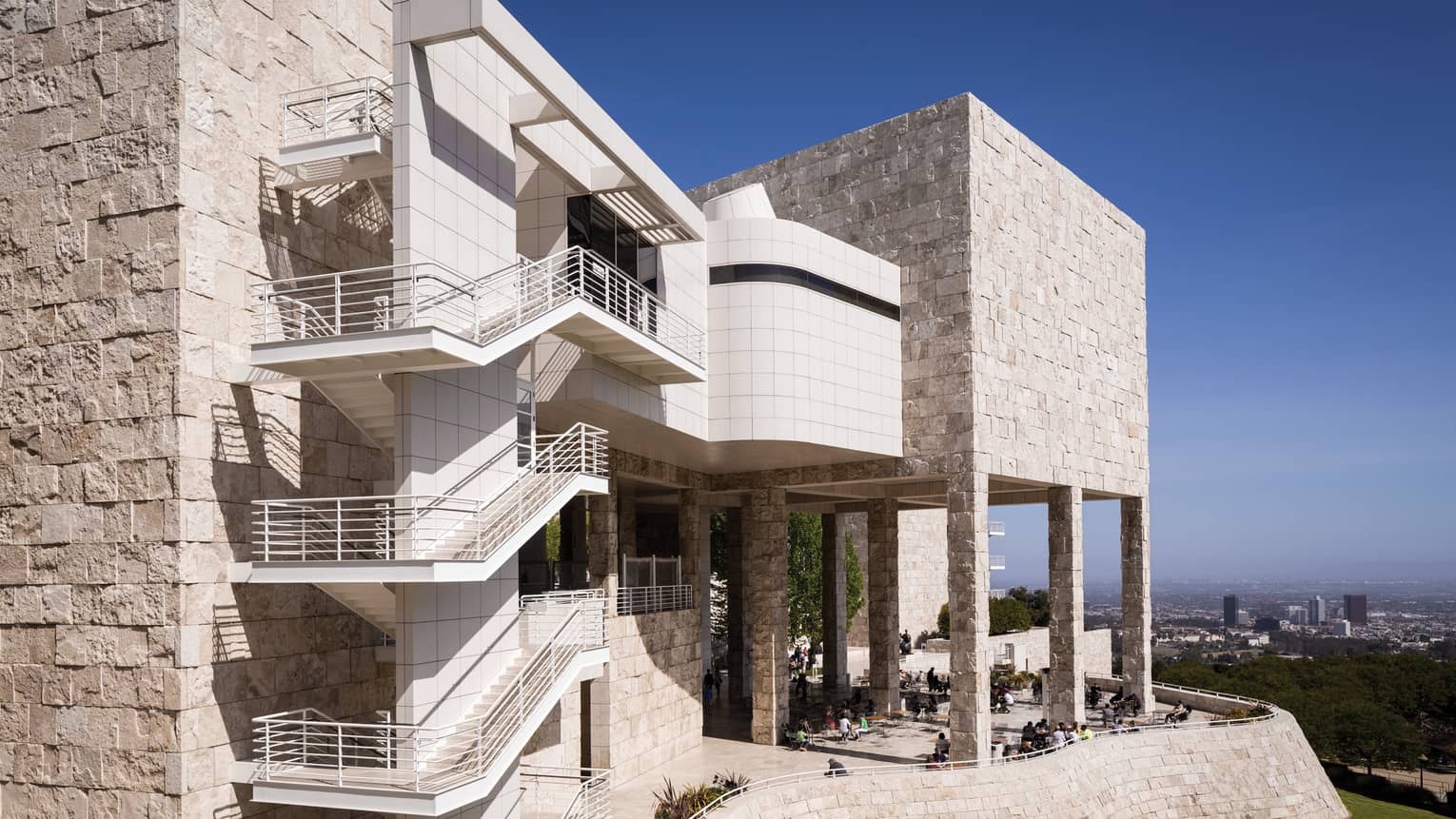 Outdoor stairways line the wall of a tan-coloured stone building with a patio full of people, a city skyline visible beyond.