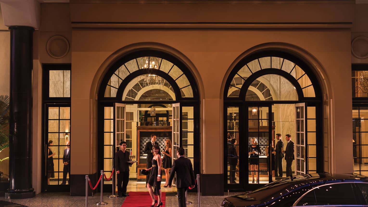 Party entrance, smiling woman wearing black cocktail dress stands on red carpet under arched doorways