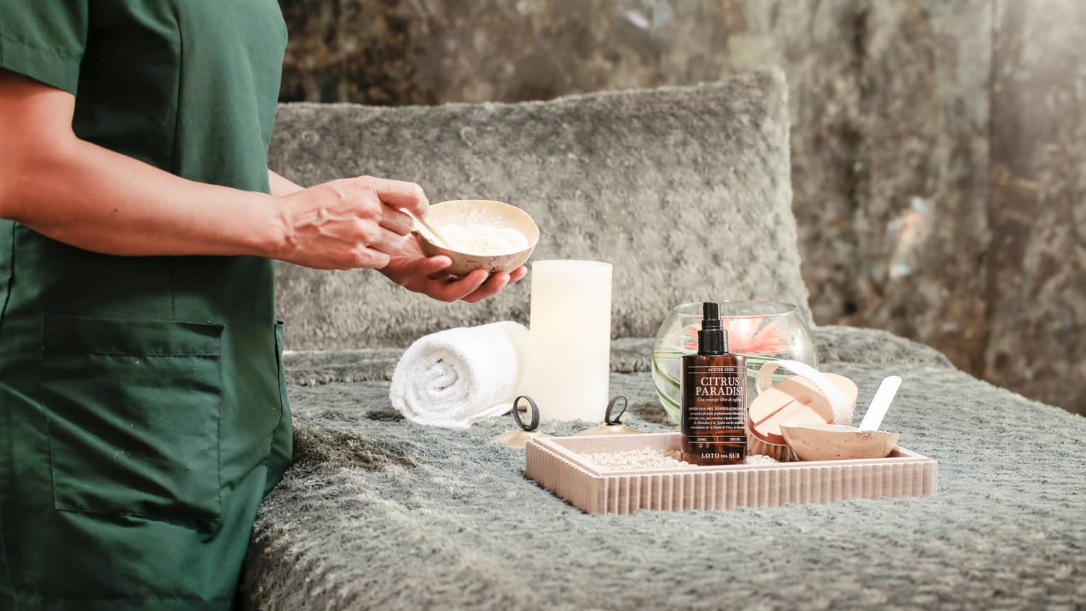 Body view of spa attendant mixing salt scrub in small bowl over bed with tray, towels, lotion