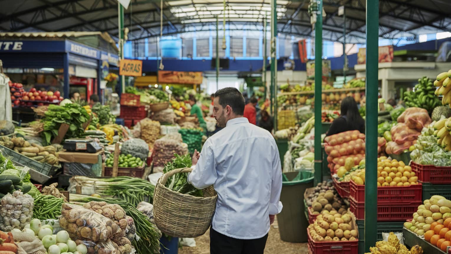 Back of man with beard carrying wicker basket through fruit market aisle, produce stacked on shelves