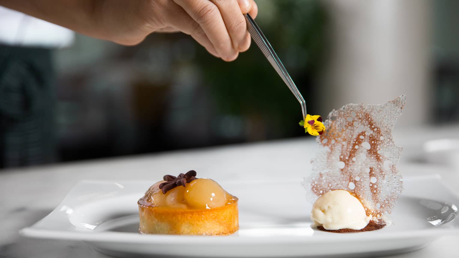 Hand with tweezers placing small yellow flower on dessert garnish on plate