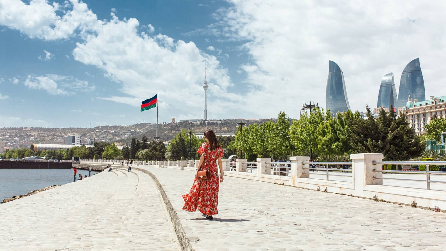 Woman wearing red dress walks along stone waterfront, skyscrapers in background