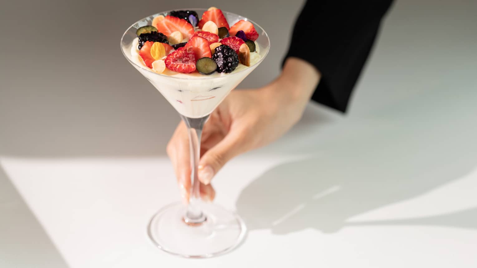 A hand delicately holds a sunlit martini glass filled with yogurt topped with summer fruits, chopped nuts, and flower petals.