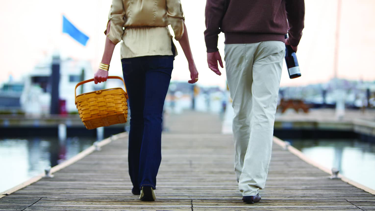 Lower half of two people walking on dock, carrying picnic basket and wine bottle