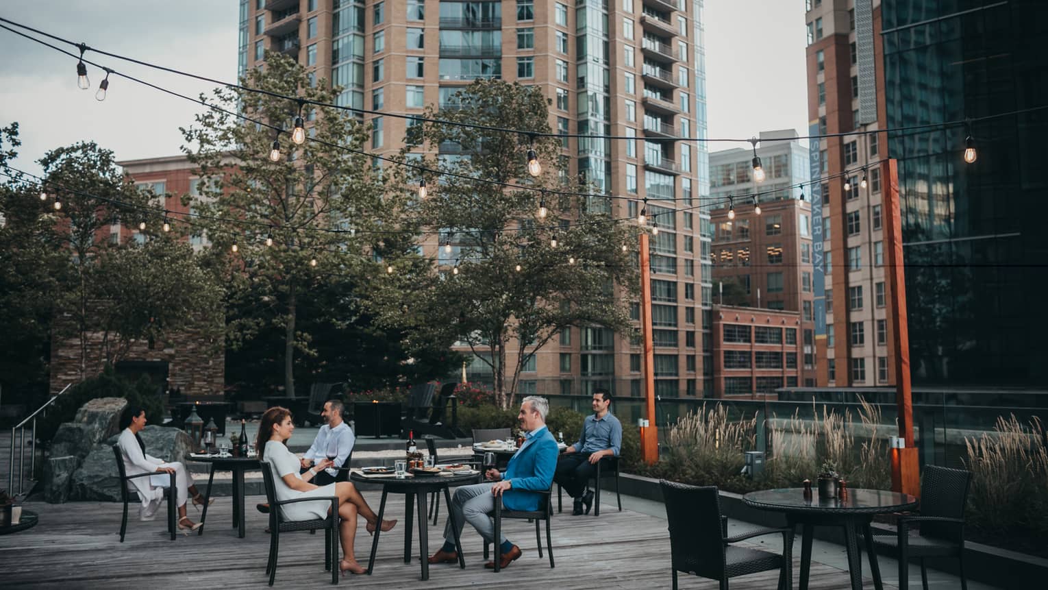 Three tables outside with a man and woman sitting at each enjoying a meal in the evening, there are trees and tall buildings in the background.