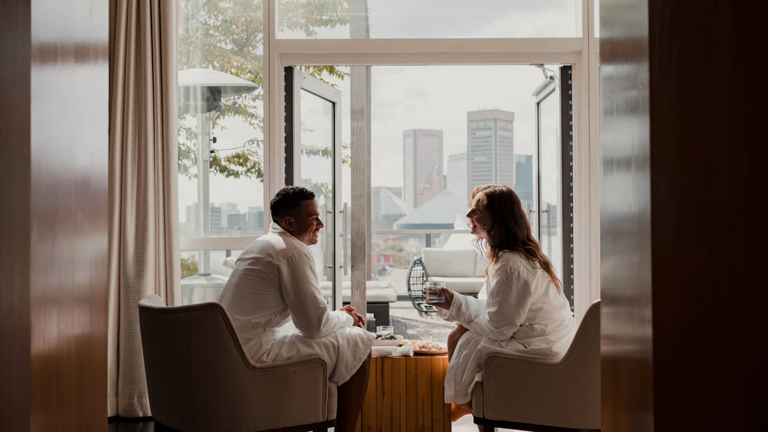 Two people in white robes sit in armchairs by an open balcony door with a cityscape view in the background, sharing a relaxed, casual conversation. 