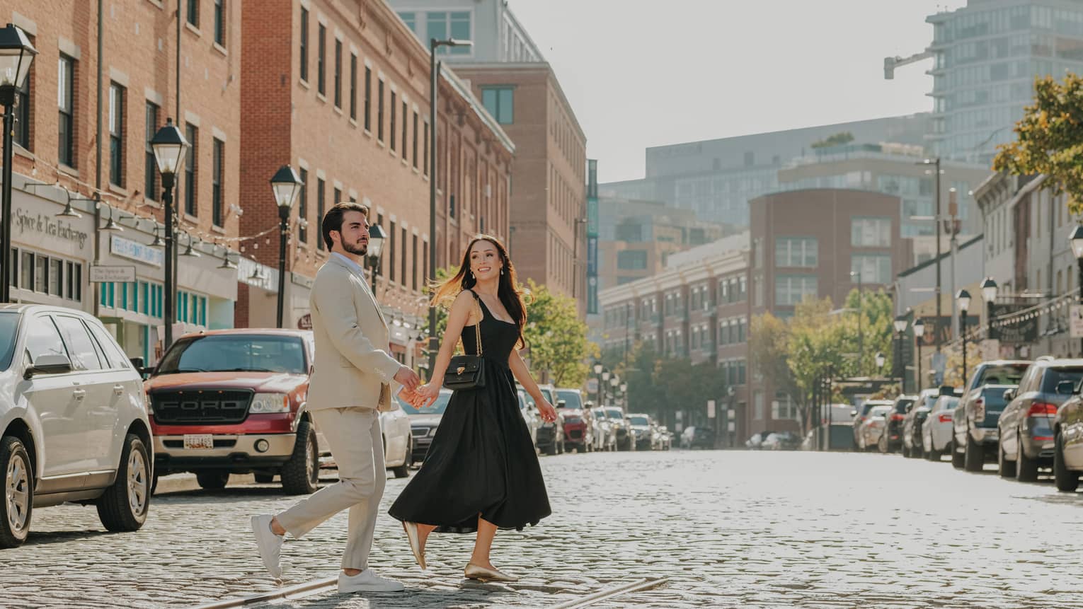 A smiling, well-dressed couple cross a cobbled street lined with stores in red-brick buildings, streetlights and parked cars.