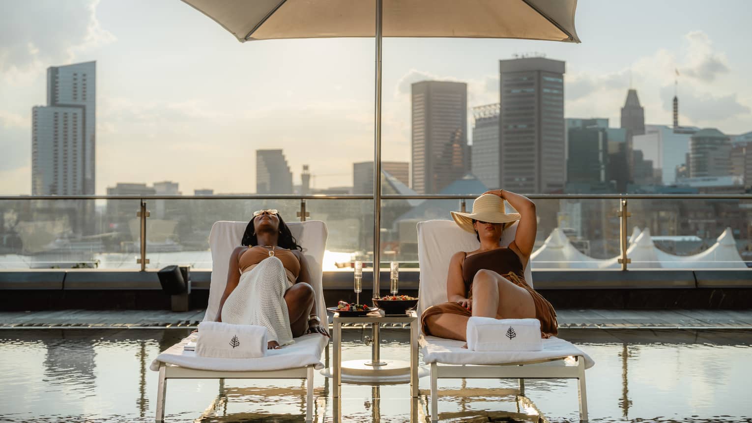 Two people recline on lounge chairs under a sun umbrella over a rooftop pool, with a background of city skyline views.