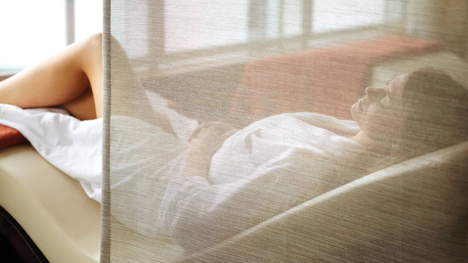 Woman wearing white bathrobe lies on curved spa chair behind sheer curtain