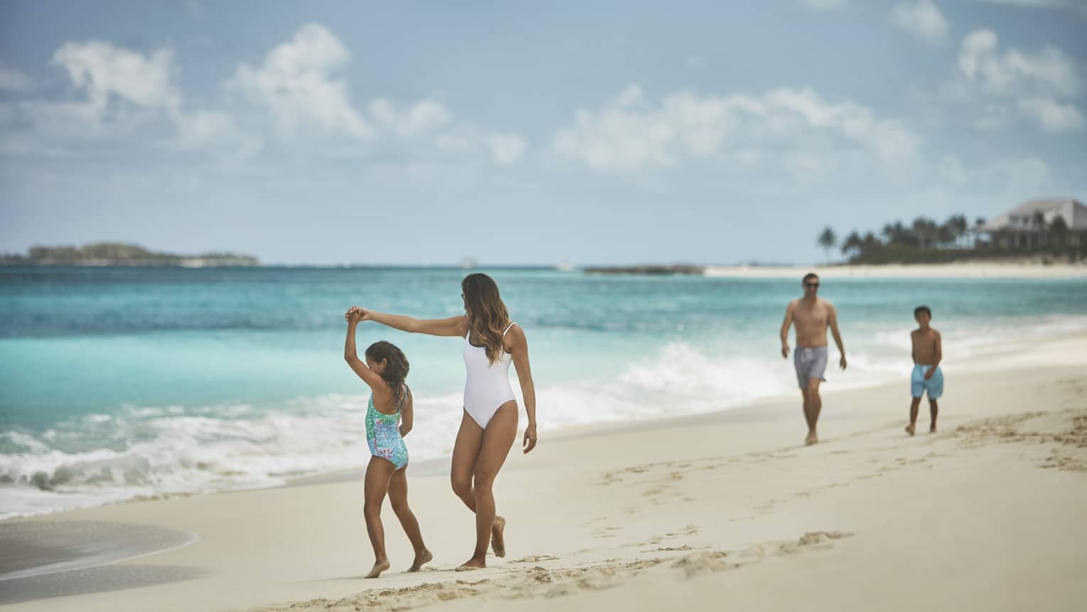 Family enjoying a walk along a sandy beach with turquoise waters, one adult and child holding hands and two others walking behind them