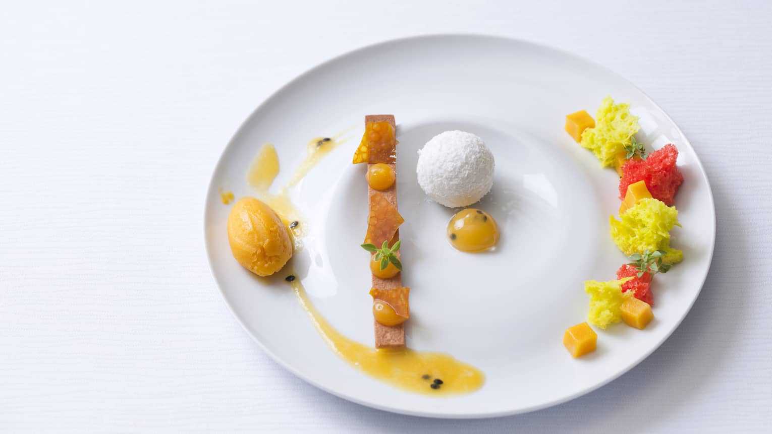 Vanilla sable, passion fruit sauce, coconut mousse ball with passion cremeux insert, marinated fresh mango in sage syrup and mango sorbet on a white plate