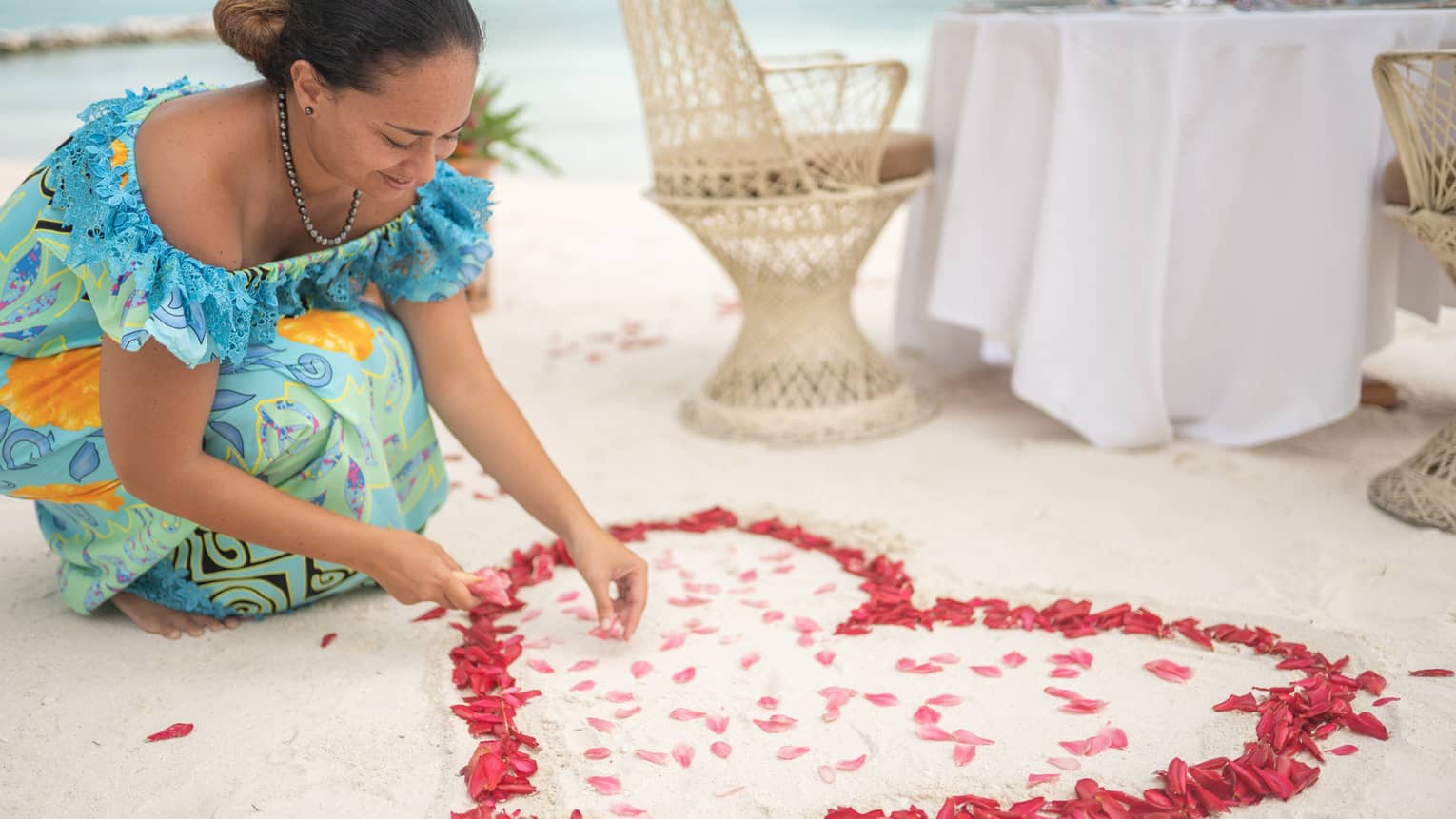 Woman arranges red rose petals into heart shape on sand beach