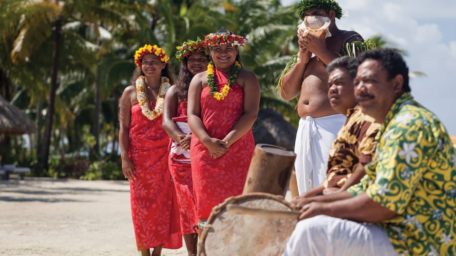 Group of people in traditional attire with leis and a conch shell, standing on a beach