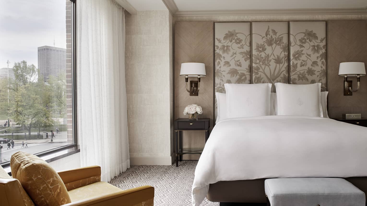 Front view of Executive Suite bed with white linens, beige fabric floral headboard, large window with white curtains, yellow armchair