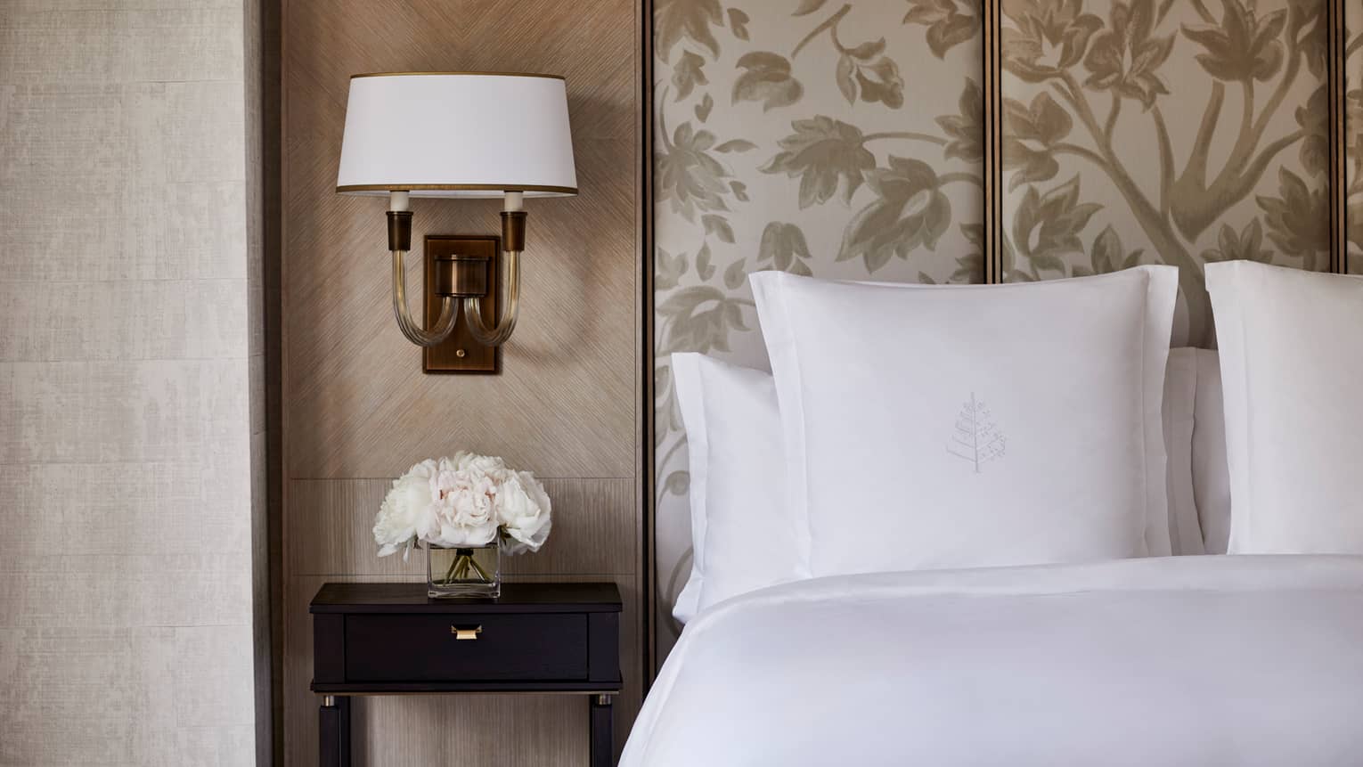 Deluxe Accessible Room close-up of bed, white pillows, beige headboard, small reading lamp on wall, white flowers on nightstand