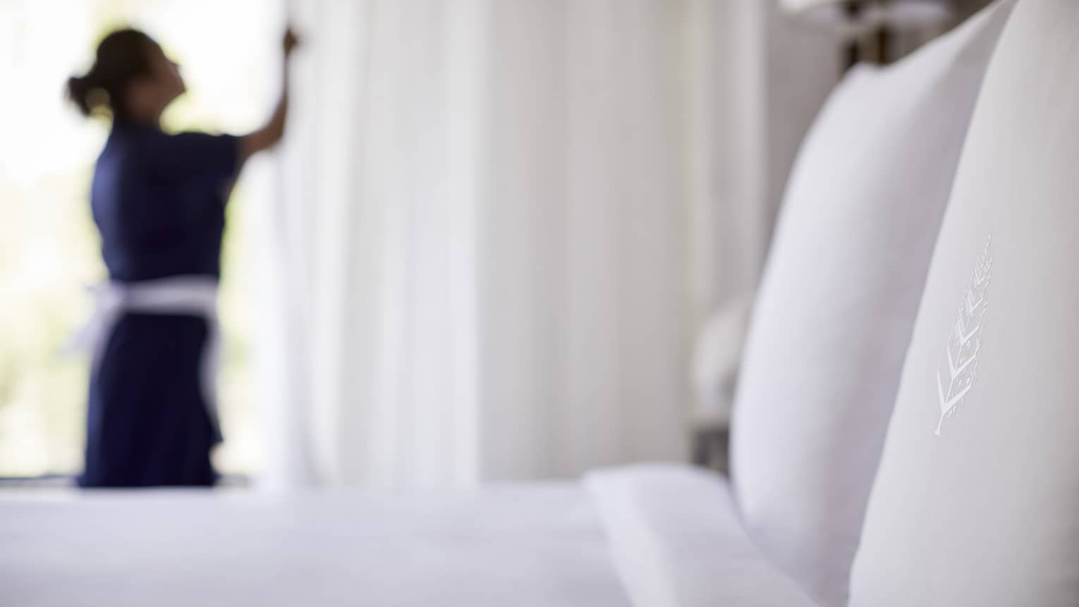 Close-up of bed, pillow with silhouette of hotel staff in uniform closing white curtains in background