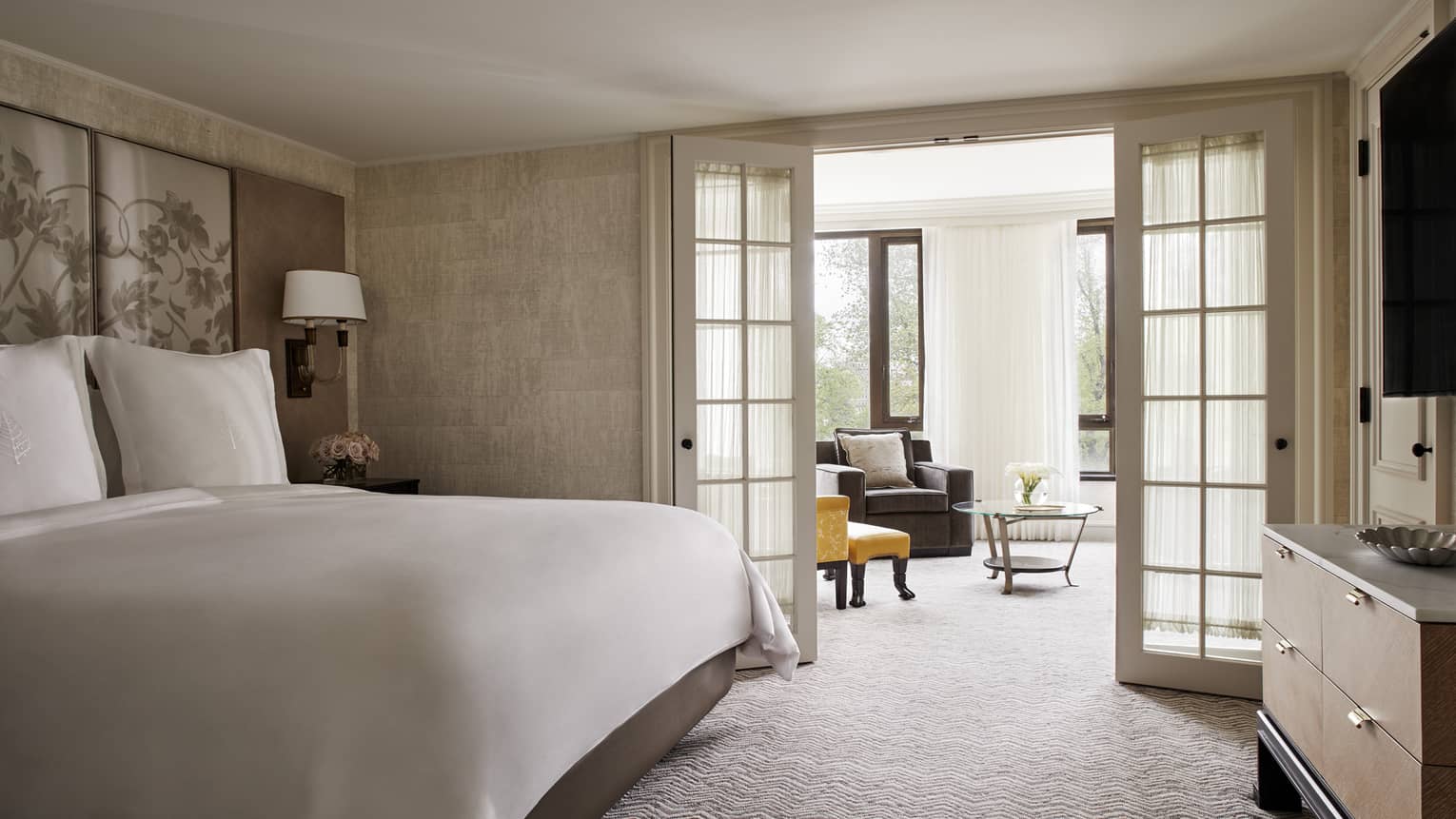 Senator Suite bed with white linens beside glass French doors leading to sunny living room with armchairs 