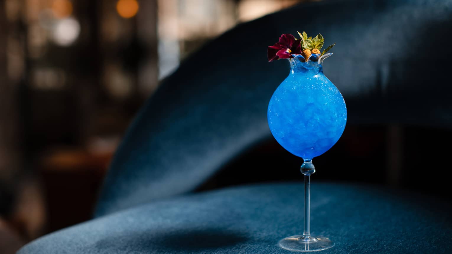 Brightly coloured cobalt blue cocktail in tall round glass and garnished with flowers