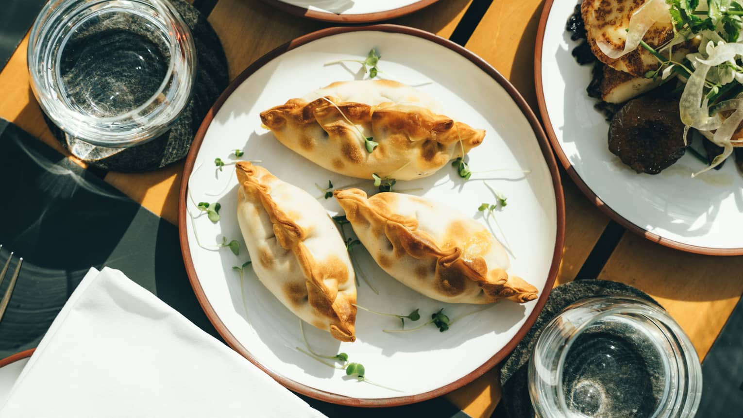 Three plump, golden-brown empanadas on a porcelain dish surrounded by sunlit water glasses and other plates of fine foods. 