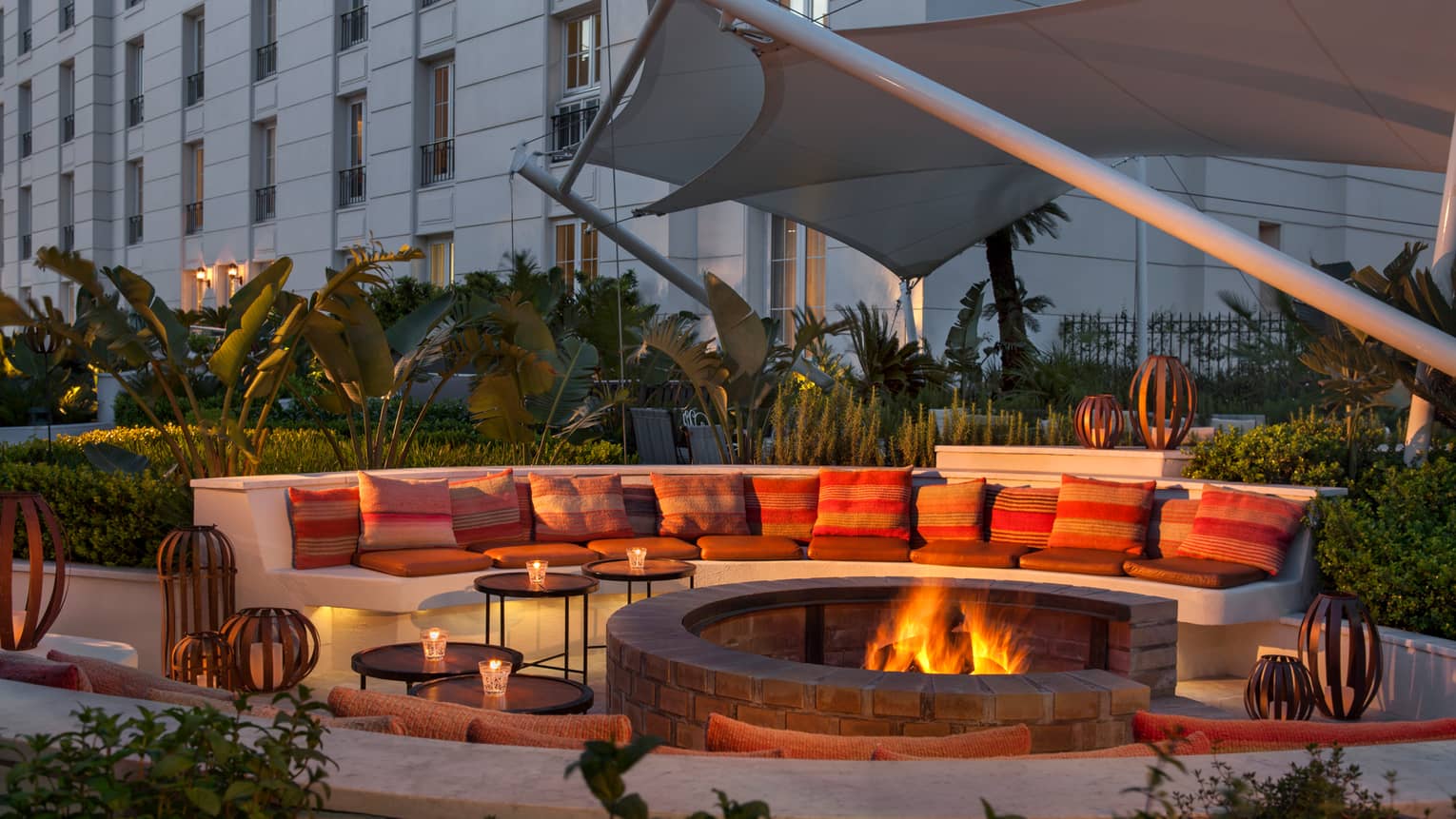 Large curved white bench with cushions around outdoor fire pit with flames at dusk