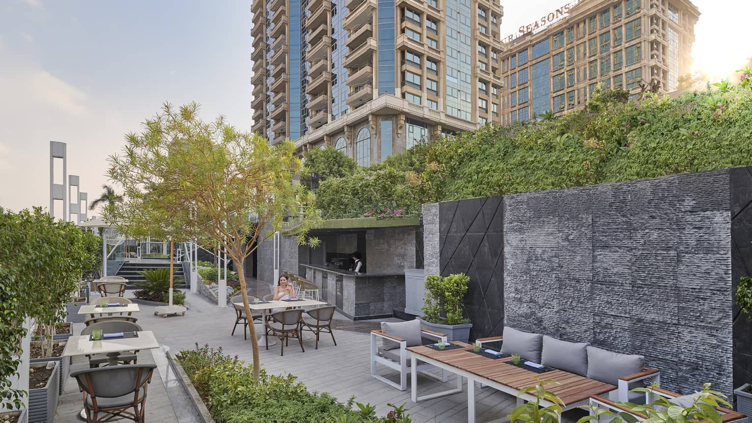 Woman seated at table on Riva restaurant concrete terrace amid greenery, hotel tower in backdrop