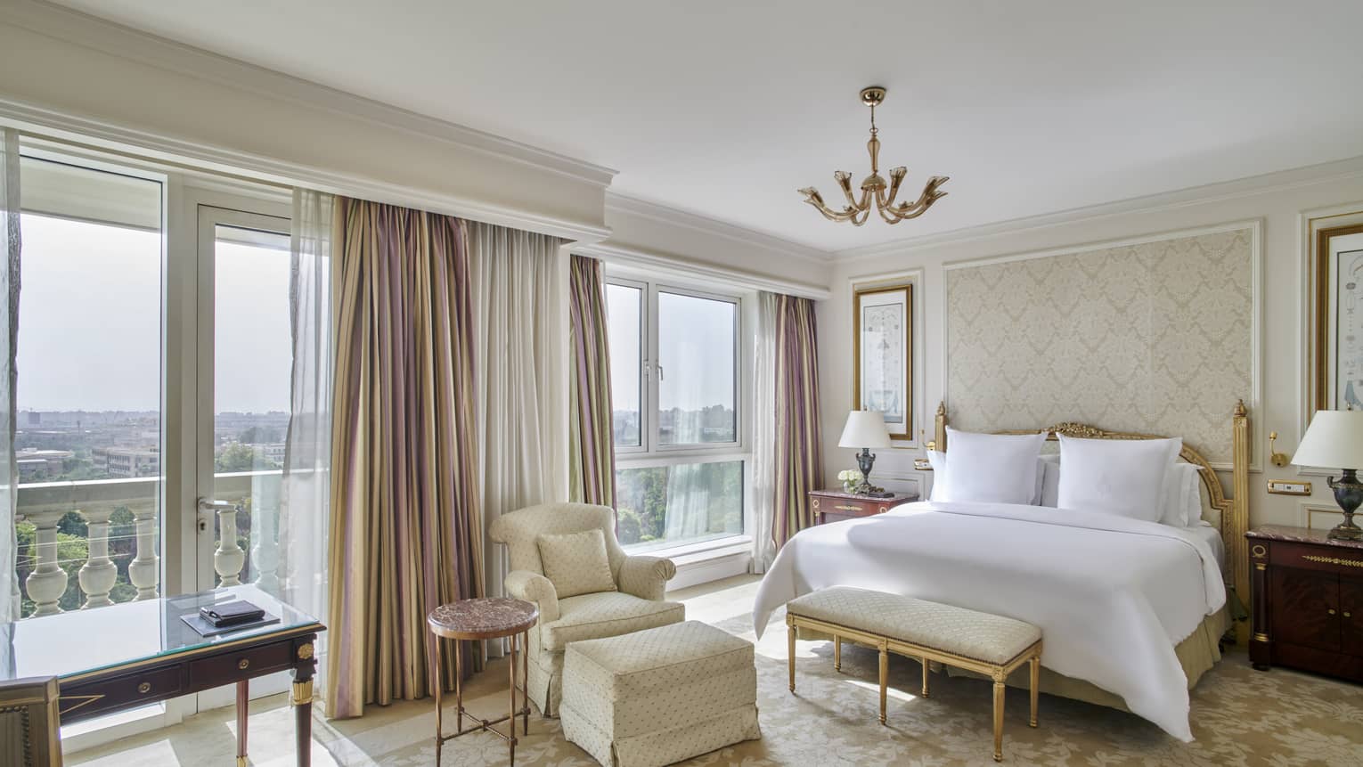 Guest room with king bed and white linens, foot bench, arm chair, floor-to-ceiling windows with city views