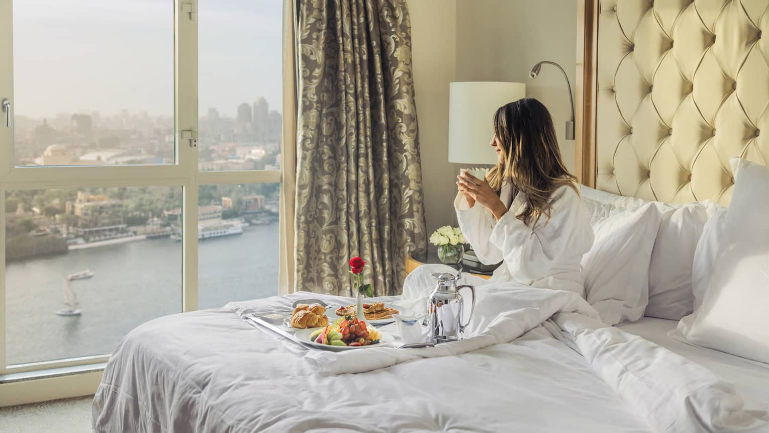 Woman sitting on a bed with a tray of food, looking toward the Nile River and Cairo from the window