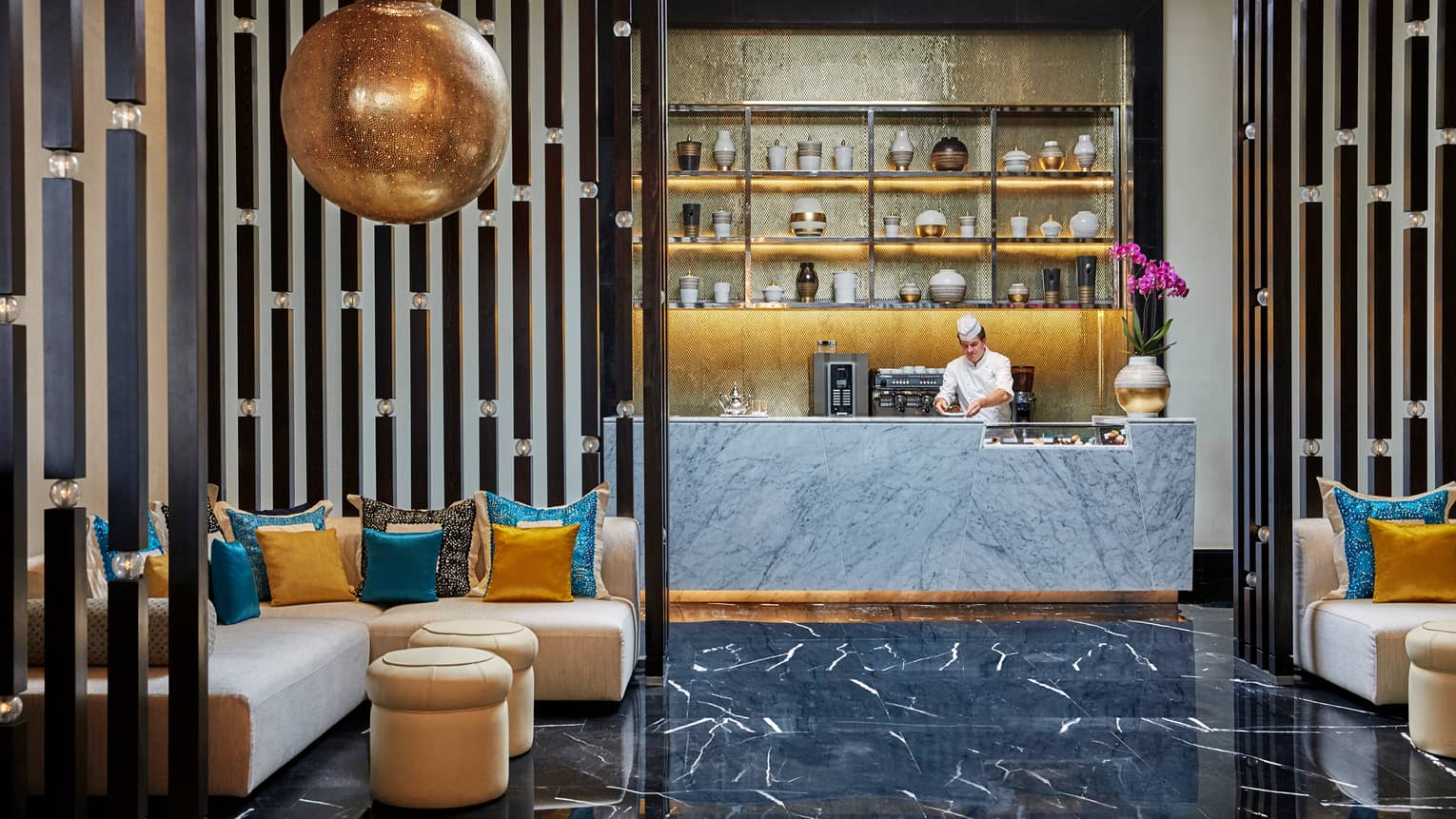 Chef behind counter, Mint lounge area with large brass hanging lamp, L-shaped sofas with colourful pillows, gilded accents