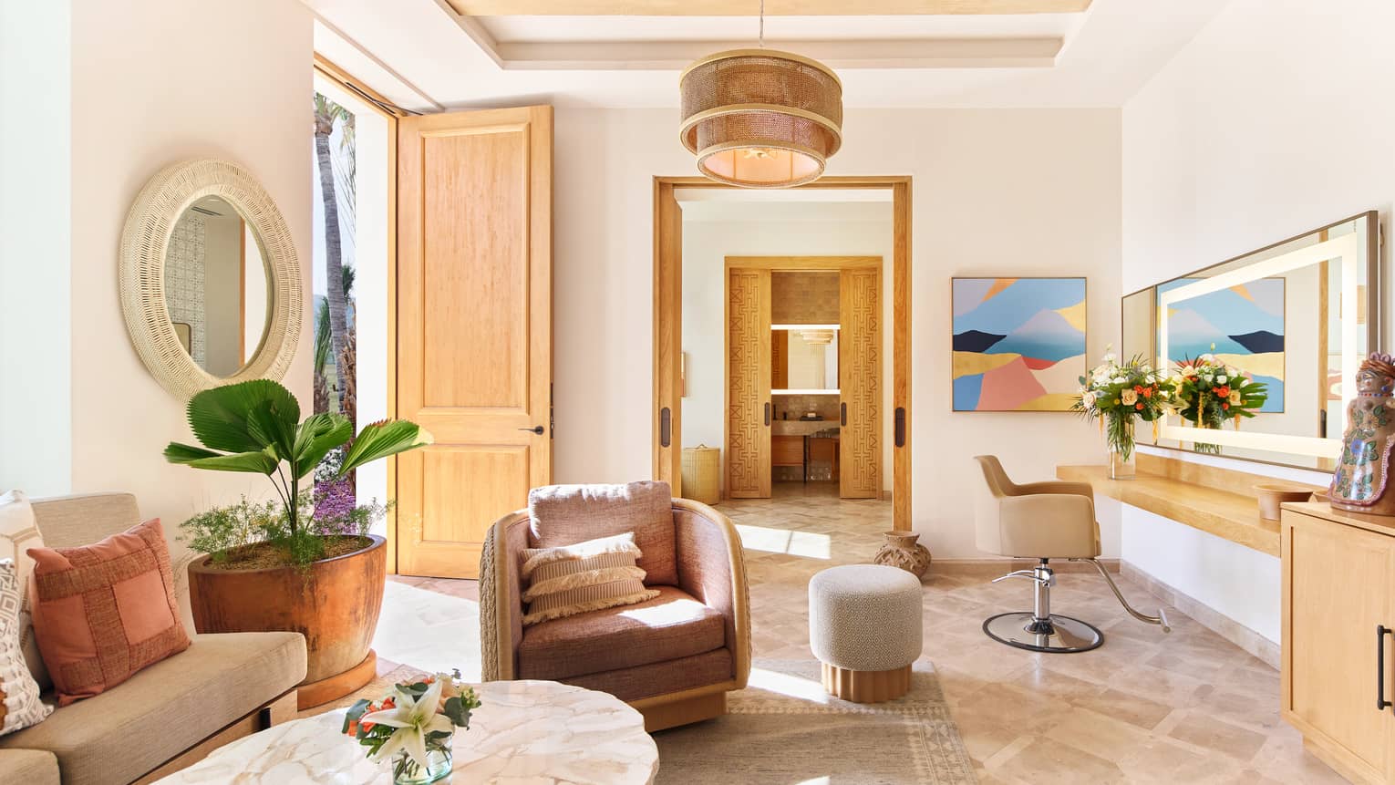 A spacious room filled with natural light, warm neutral decor, comfortable upholstered seating, artwork on the walls, floral arrangements and potted plants, and coffee and end tables