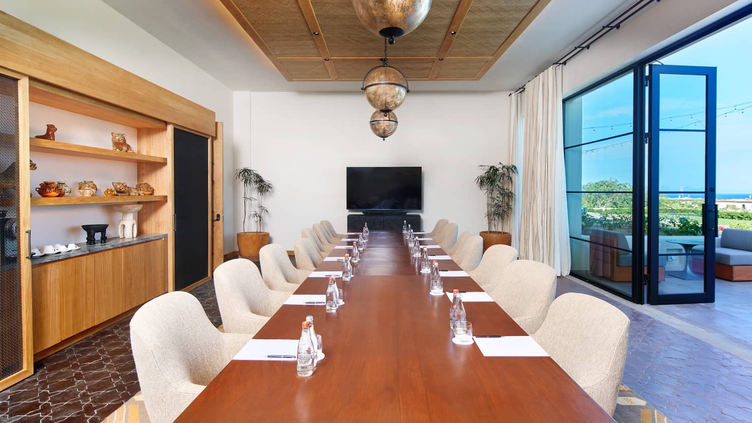 A long meeting room with long wood table, beige upholstered chairs, floor-to-ceiling windows and doors that open onto a patio, a flat-screen TV, globe-shaped pendant lighting, a sidebar with shelving above it, and overall neutral-toned decor and finishes