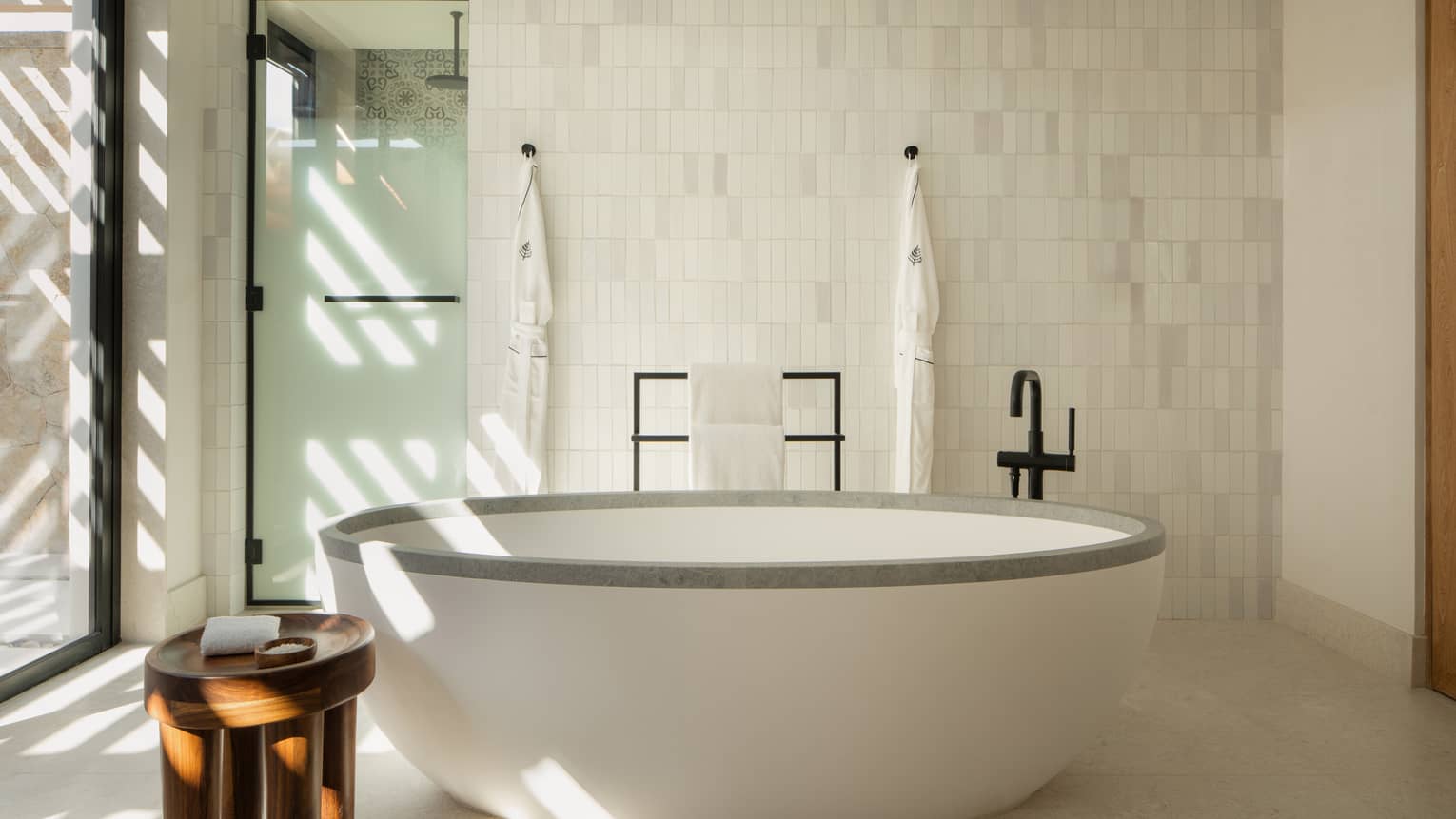 Standalone oval tub in luxury hotel bathroom, and frosted glass door leading to a separate shower. Two bathrobes hang on tiled wall's hooks.