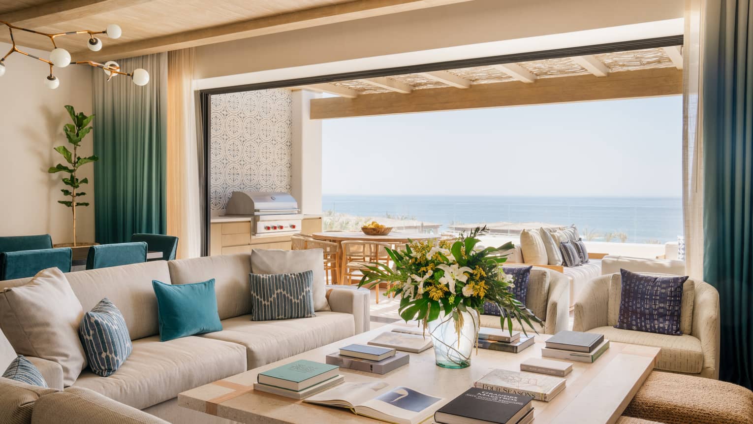 Luxury villa living room with sofa, chairs and ottomans around a large square coffee table, with a wall open to ocean-view pool terrace, at Four Seasons Resort Cabo San Lucas
