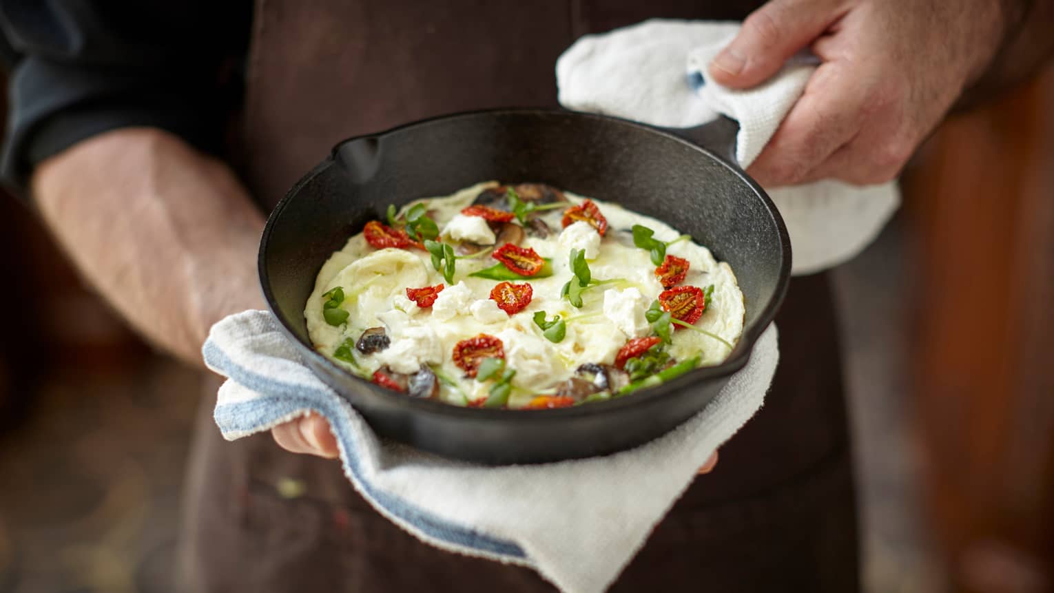 Close-up of man in black uniform holding iron pan with egg, sun dried tomatoes, feta cheese and herbs