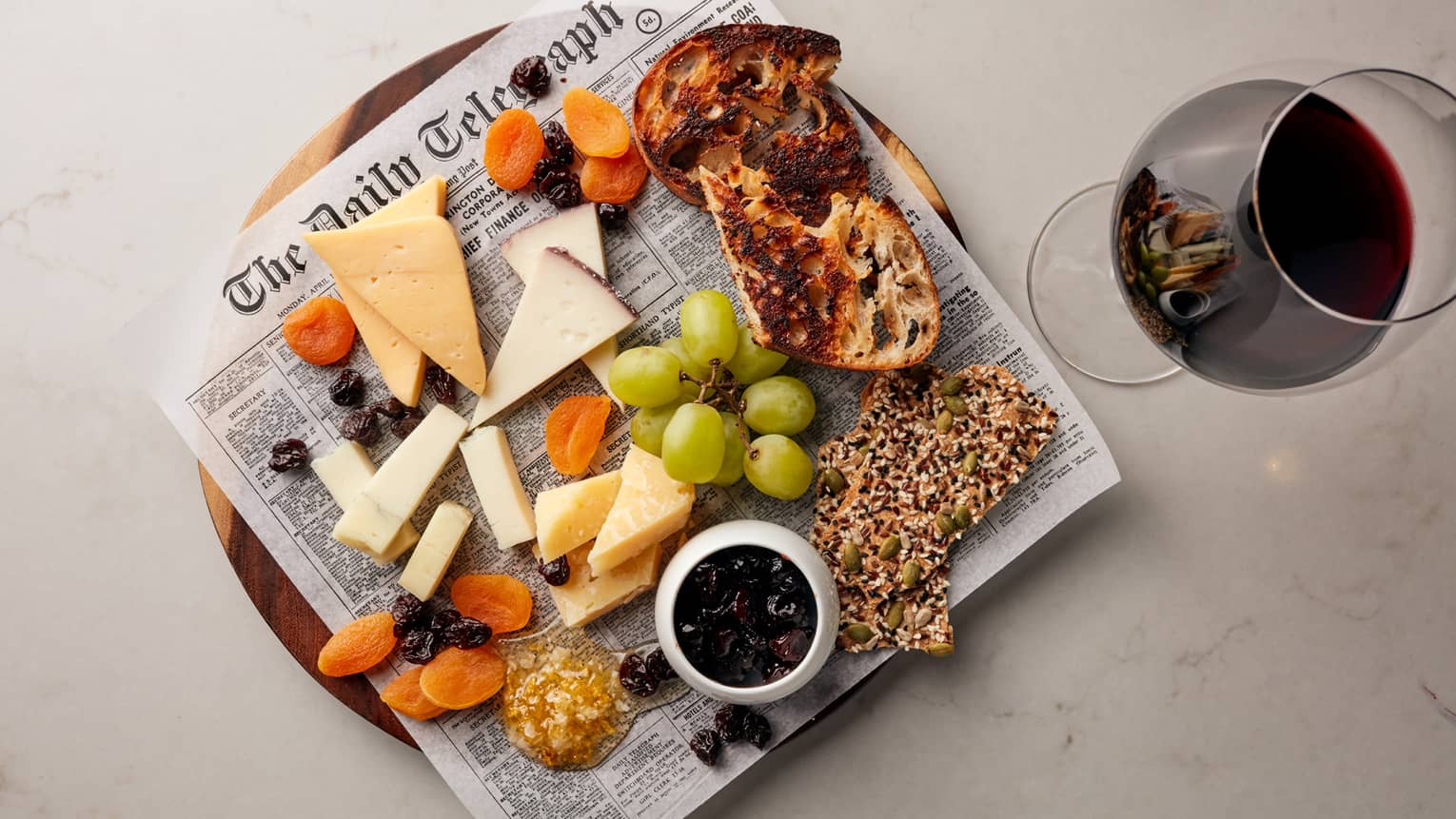 Cheese, grapes, crackers and bread on newspaper on platter next to glass of red wine