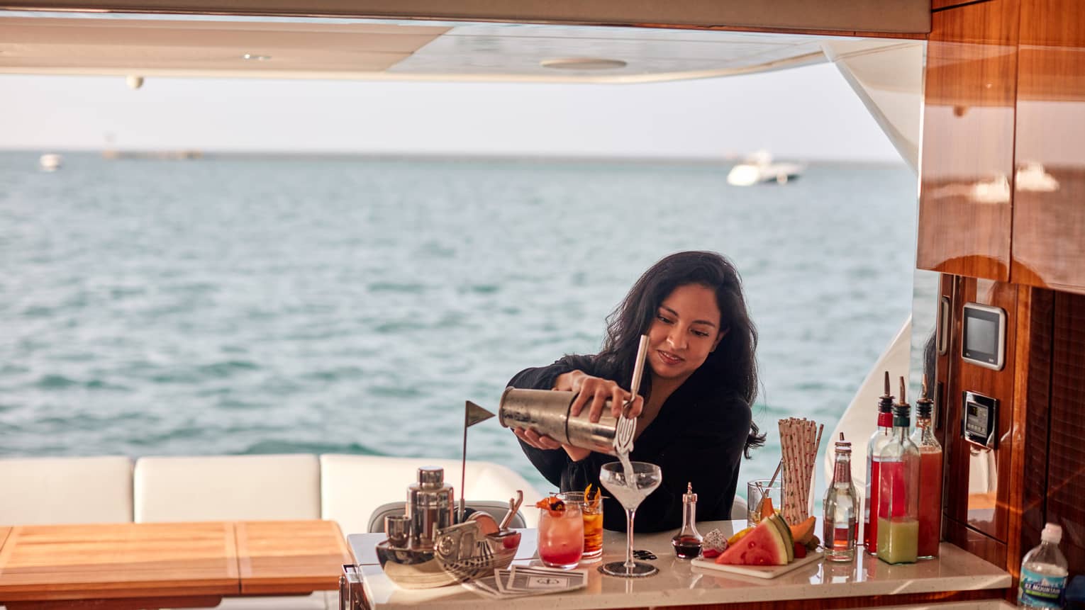 On a yacht, a passenger makes a cocktail at a counter with food and beverages on it, in the background is an ocean view.