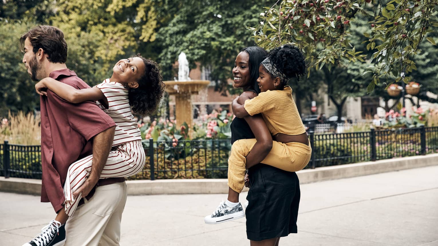Two parents each holding a child on their back walk by a city fountain