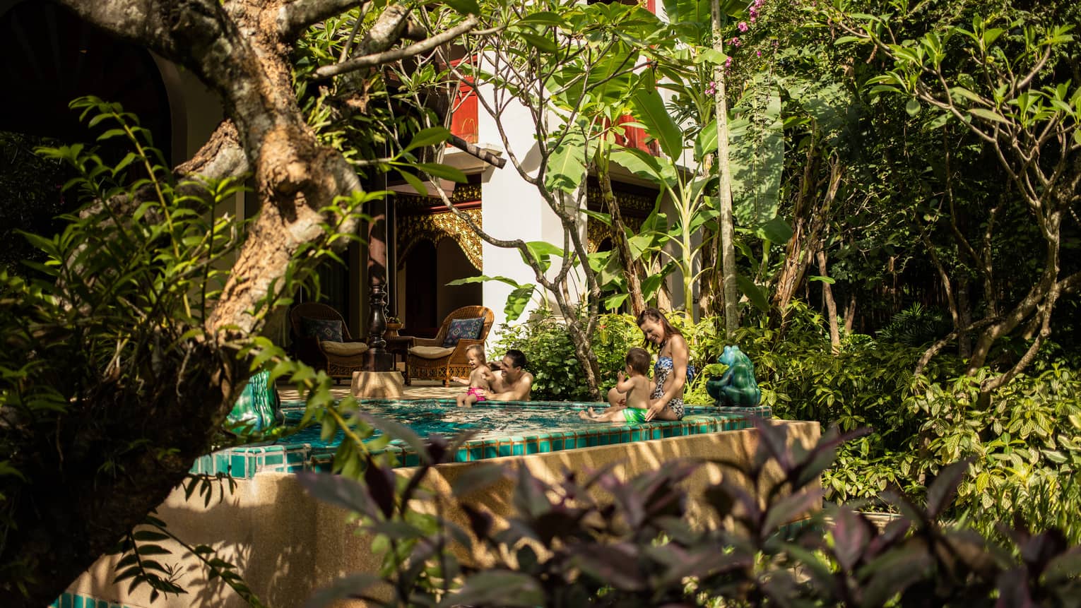 Couple with two babies sit on edge of private pool in front of bungalow by garden