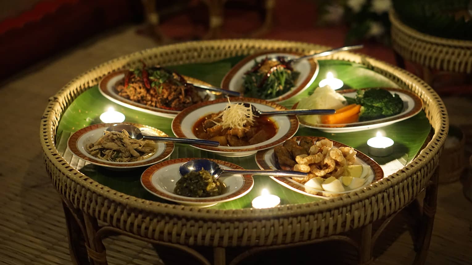 A round wicker tray laden with several meat and vegetable delicacies on a leaf-coloured mat and punctuated with tealights.