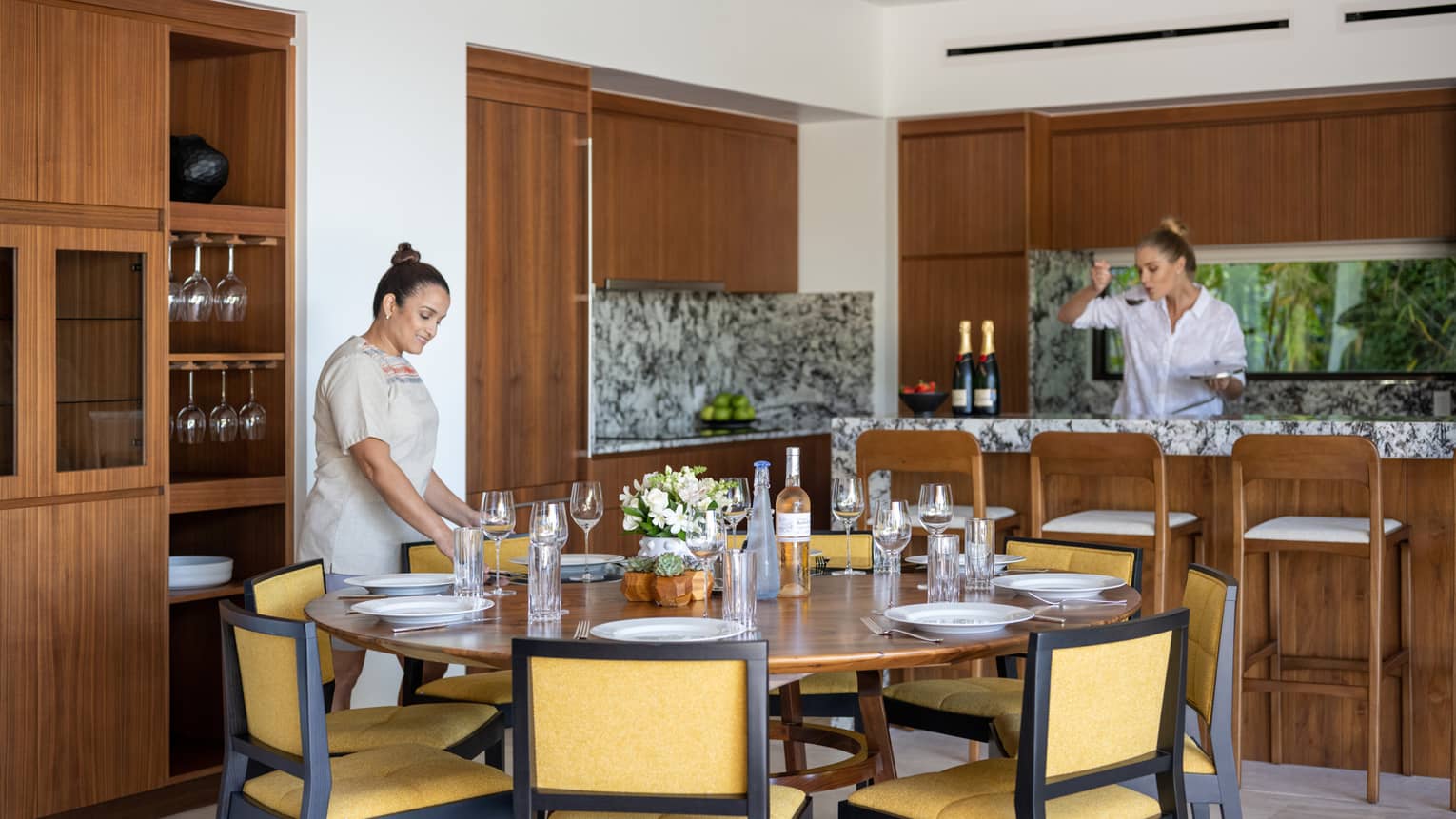 Woman setting round dining table and woman at kitchen counter