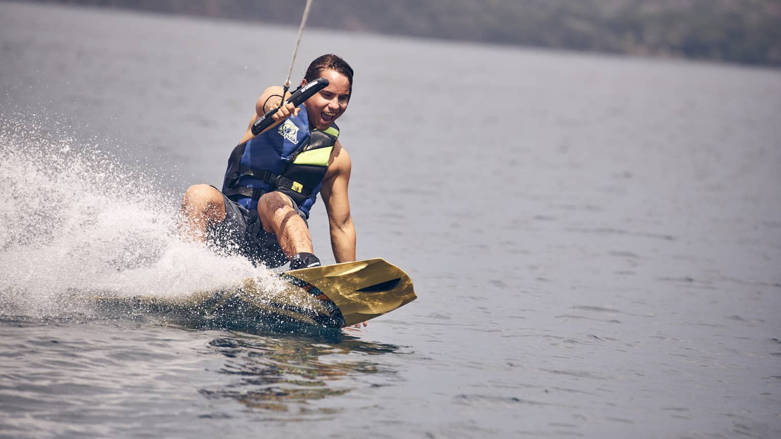 Young man wearing a blue and green life vest and blue swim trunks wakeboarding through the water